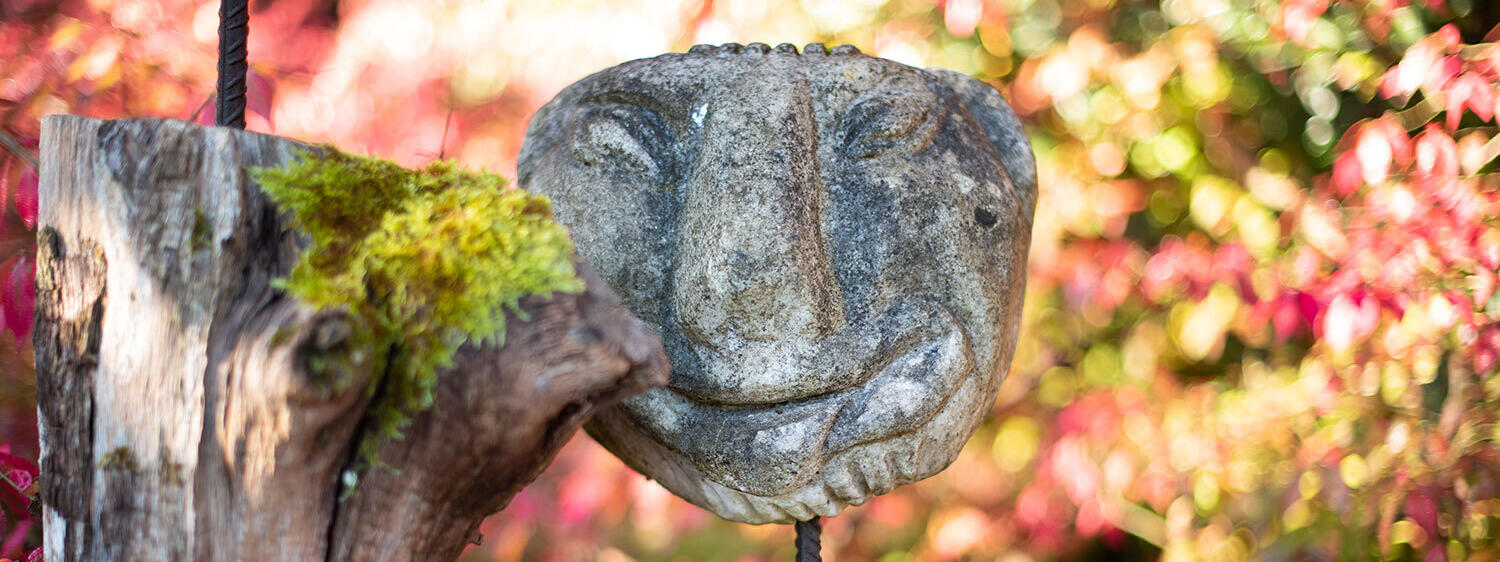 Eine lächelnde Steinskulptur umgeben von herbstlichen roten Blättern und Moos auf Holz im ZIST Garten.