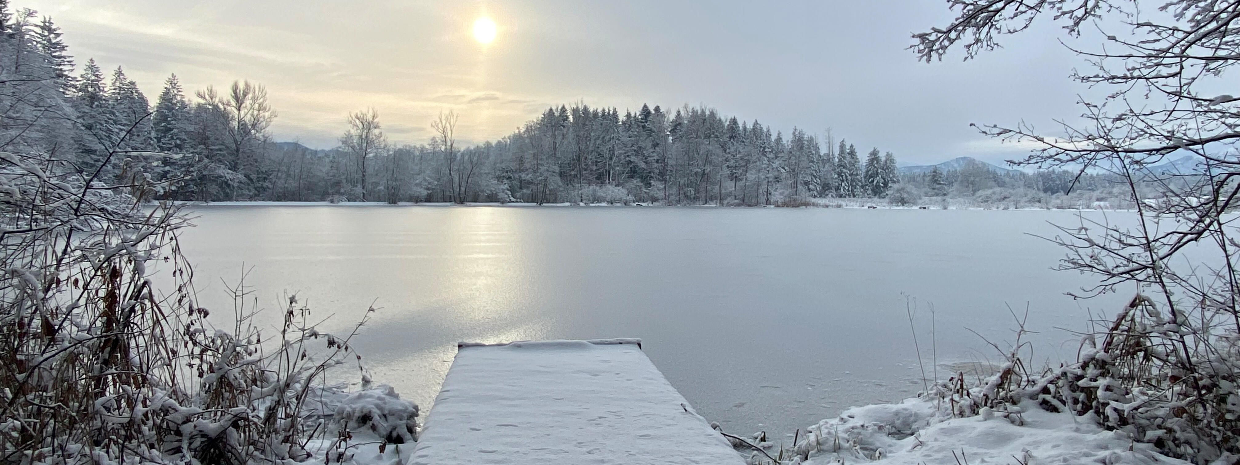 Schneebedeckter Steg, der zu einem gefrorenen Teich führt, mit der Sonne, die über der winterlichen Landschaft aufgeht.