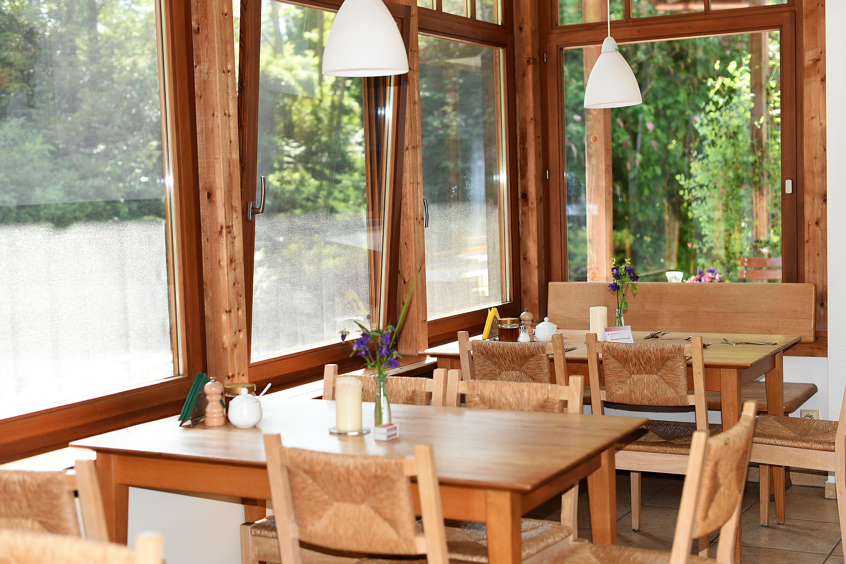 Speiseraum mit Holztischen und Stühlen vor großen Fenstern und Blick in den Garten
