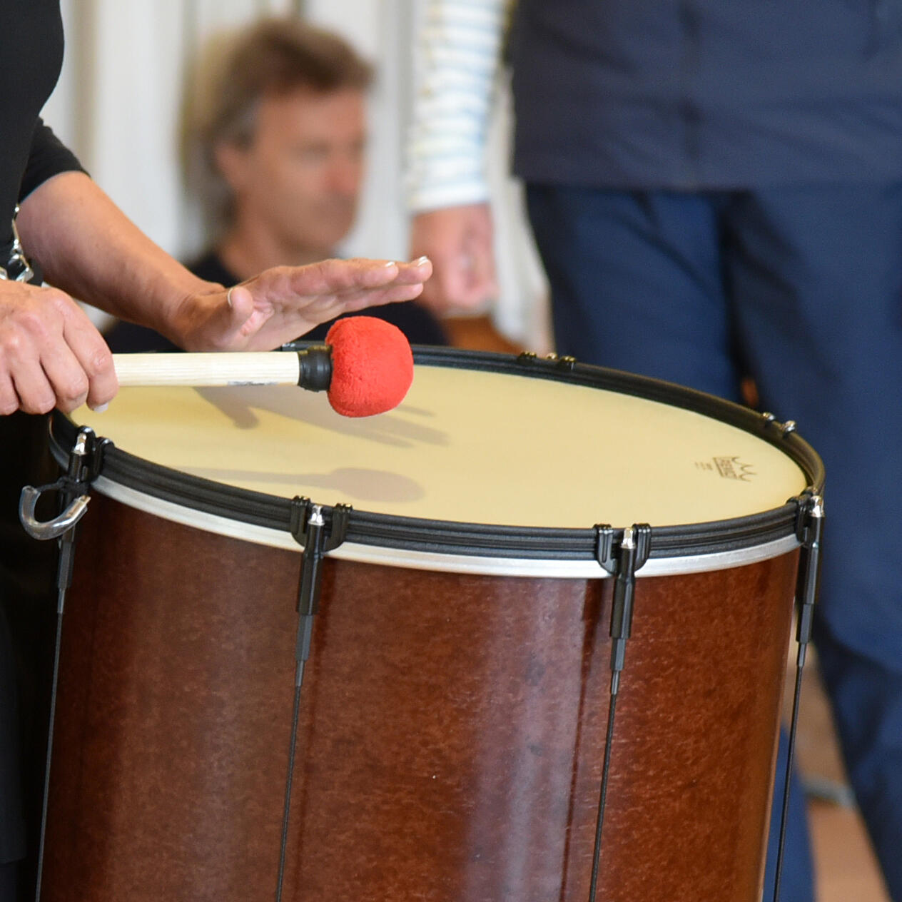 TaKeTiNa Rhythmusarbeit - Eine Person schlägt mit einem roten Schlägel auf eine große Trommel, während im Hintergrund weitere Menschen anwesend sind.