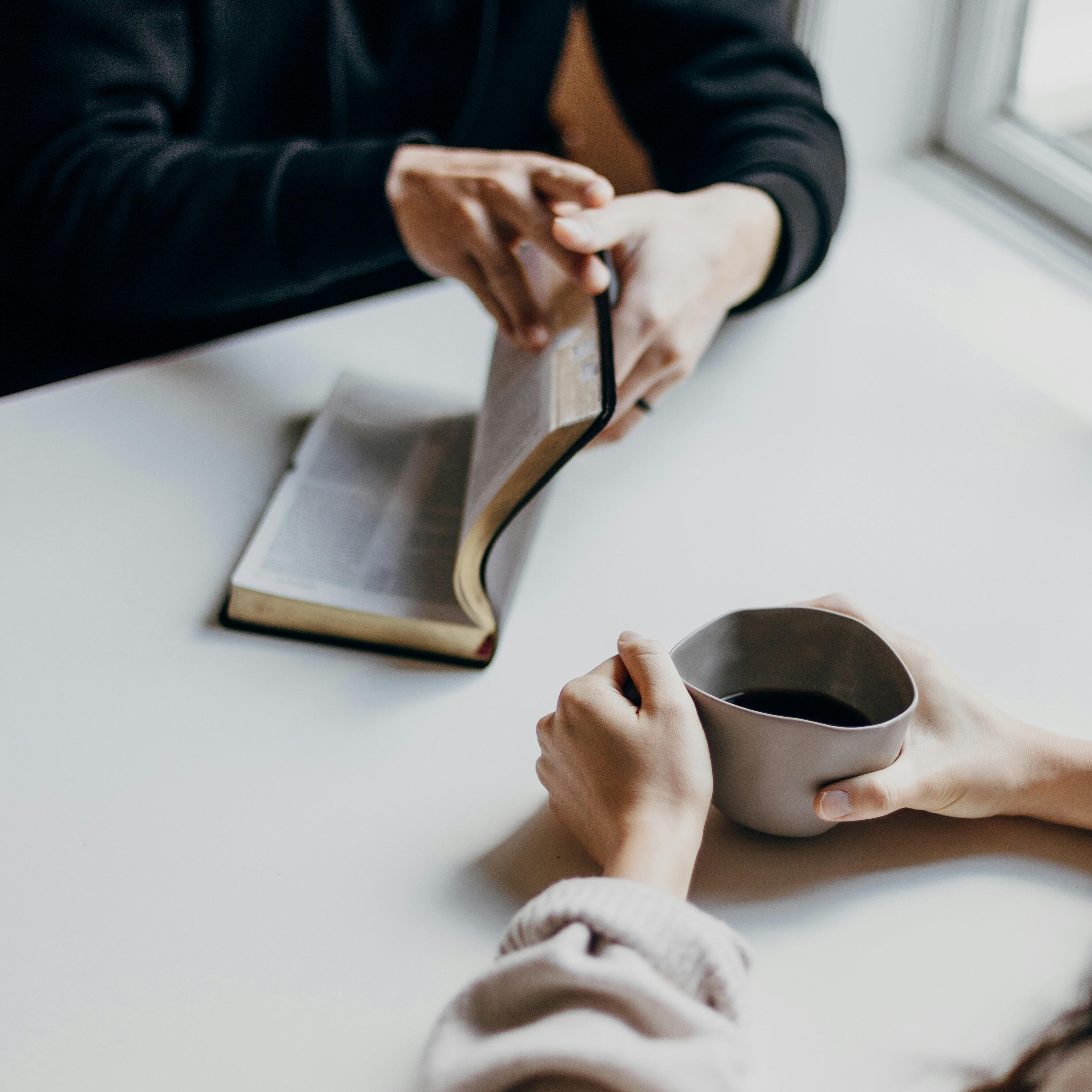 Zwei Personen sitzen an einem Tisch, eine hält eine Kaffeetasse, während die andere eine Seite in einem aufgeschlagenen Buch umblättert. Die Szene strahlt Ruhe, Nachdenklichkeit und Verbundenheit aus.