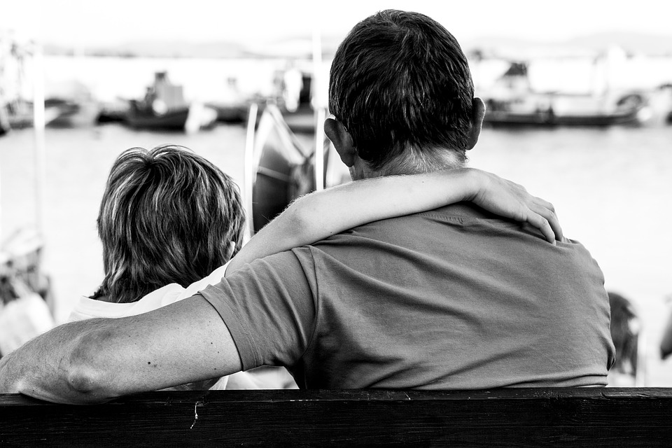 Vater und Kind sitzen auf der Bank, das Kind hat den Arm um die Schultern des Vaters gelegt, der Vater den Arm um das Kind. Ansicht von hinten, schwarz-weiß Foto. Sie schauen gemeinsam auf eine Szene in einem Bootshafen